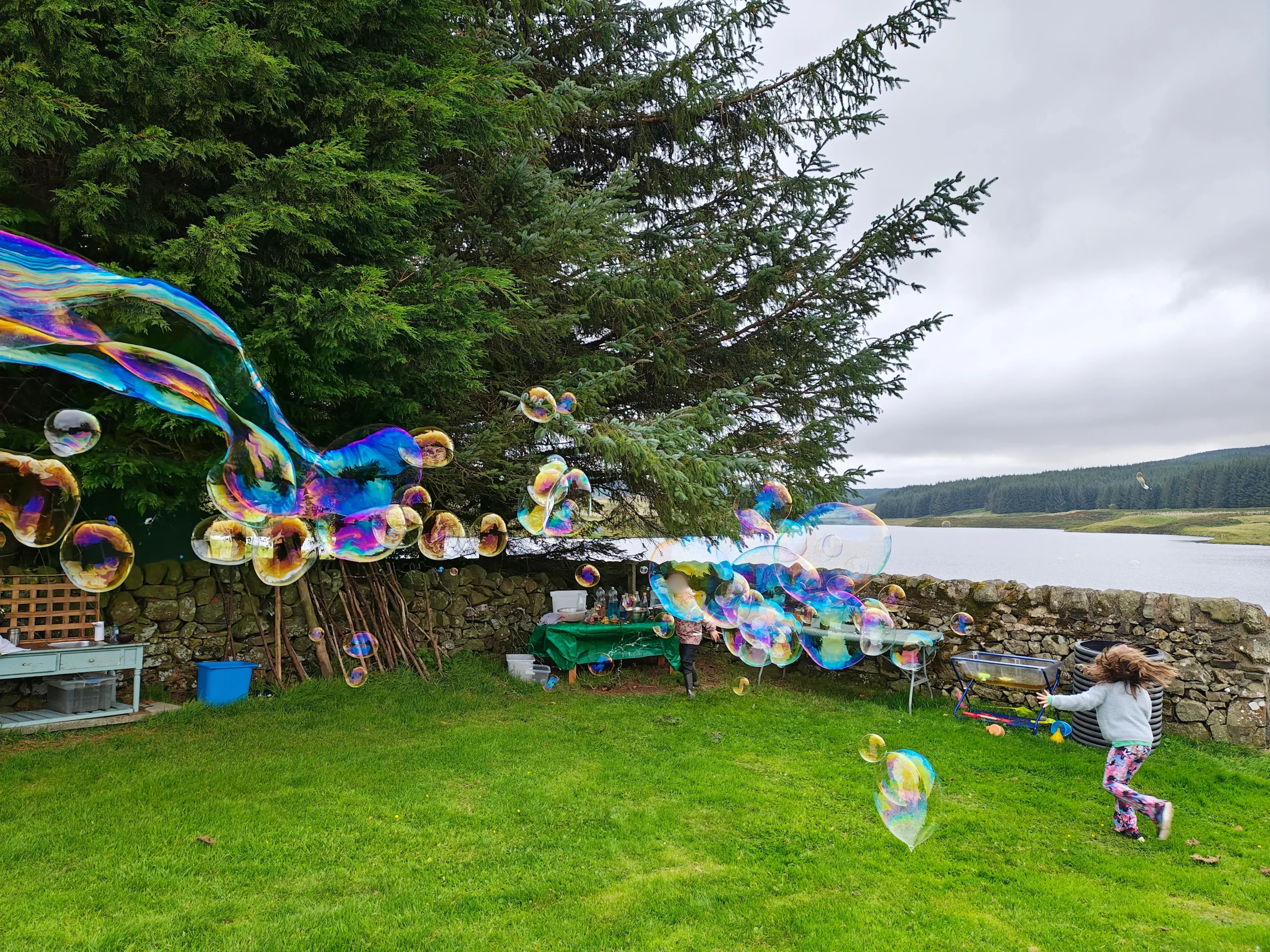 Forest school outdoor learning: children chase giant bubbles by a lake, nature-based play for wellbeing in a small group.