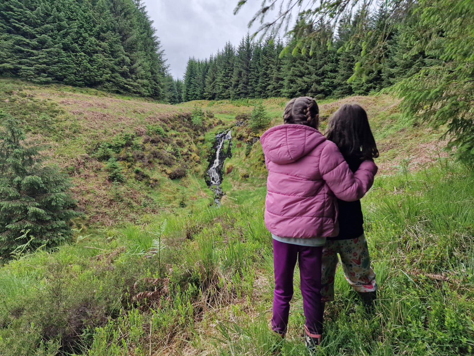 Two children in forest school pause by a waterfall, nature-based play boosting wellbeing and small-group outdoor learning.